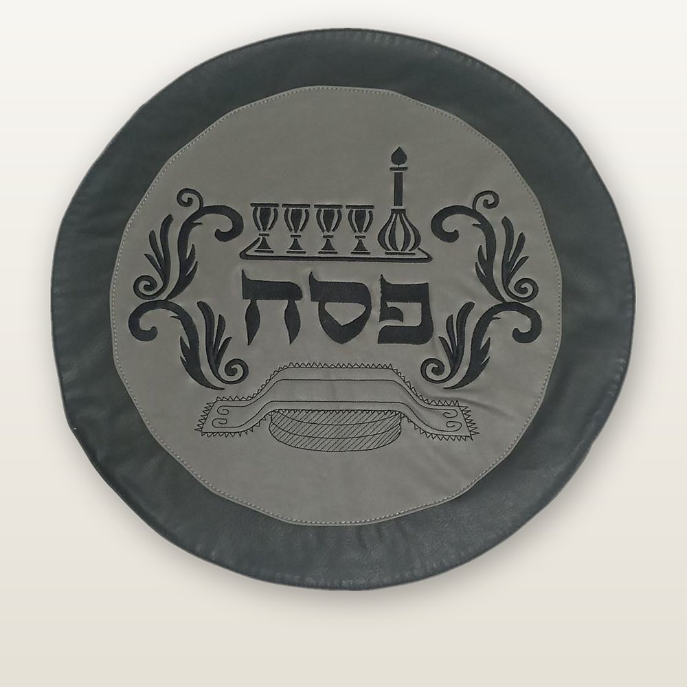 כיסוי מצה עגול מעור, שוליים באפור-פחם ומרכז אפור בהיר, רקמה שחורה של ארבע כוסות, קנקן ומצה